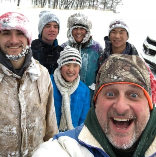 Dad taking big smiling selfie with family behind him in the snow