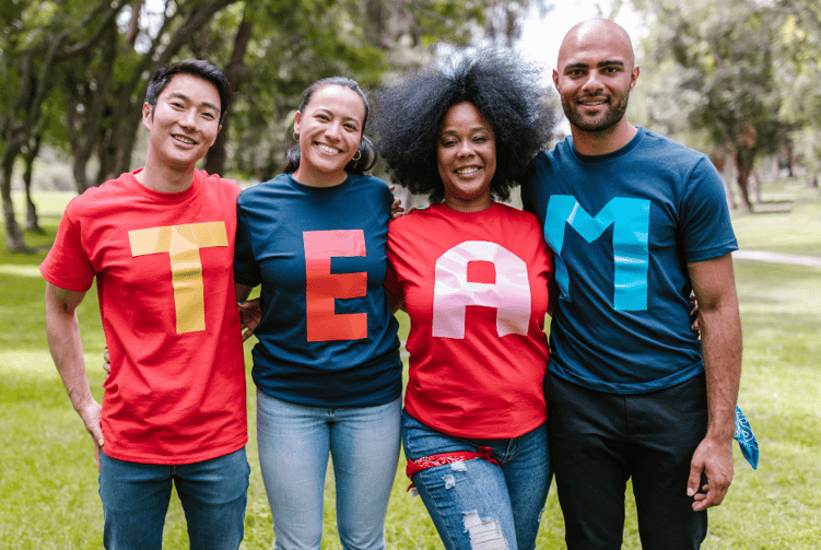 Diverse group of people with their arms around each other wearing shirts that together spell "T E A M"